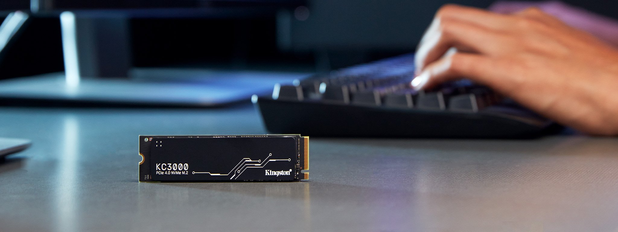 Un SSD KC3000 M.2 est posé à la verticale sur un bureau, avec des mains sur un clavier à l’arrière-plan