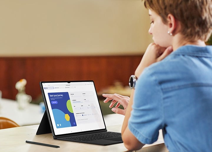 Une femme dans un caf&eacute; travaille sur une brochure &agrave; l&rsquo;aide d&rsquo;une Galaxy Tab S9 avec le clavier Book Cover Keyboard install&eacute;. Un S Pen est plac&eacute; &agrave; c&ocirc;t&eacute; de la tablette.