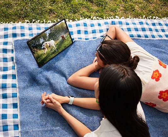 Deux femmes allong&eacute;es sur une couverture de pique-nique sur l&rsquo;herbe regardent une vid&eacute;o sur une Galaxy Tab S9 en mode Paysage avec l&rsquo;&eacute;tui Smart Book Cover install&eacute;, maintenue &agrave; l&rsquo;aide de la couverture arri&egrave;re pliable.