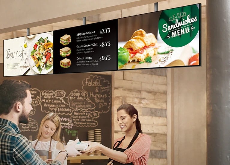 Le personnel d&rsquo;un magasin de sandwichs remet un sandwich &agrave; un client. Au-dessus d&rsquo;eux, la s&eacute;rie SM5J indiquant un panneau de menu a &eacute;t&eacute; install&eacute;e et affiche des menus avec promotions sur les brunch.