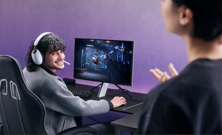 Homme portant un casque INZONE H9 assis &agrave; un bureau avec moniteur et jeu &agrave; l'&eacute;cran, parlant &agrave; un ami au premier plan &agrave; droite