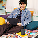 Image of a person using a tablet on a bed whilst talking into a black SRS-XB100 speaker balanced on a book