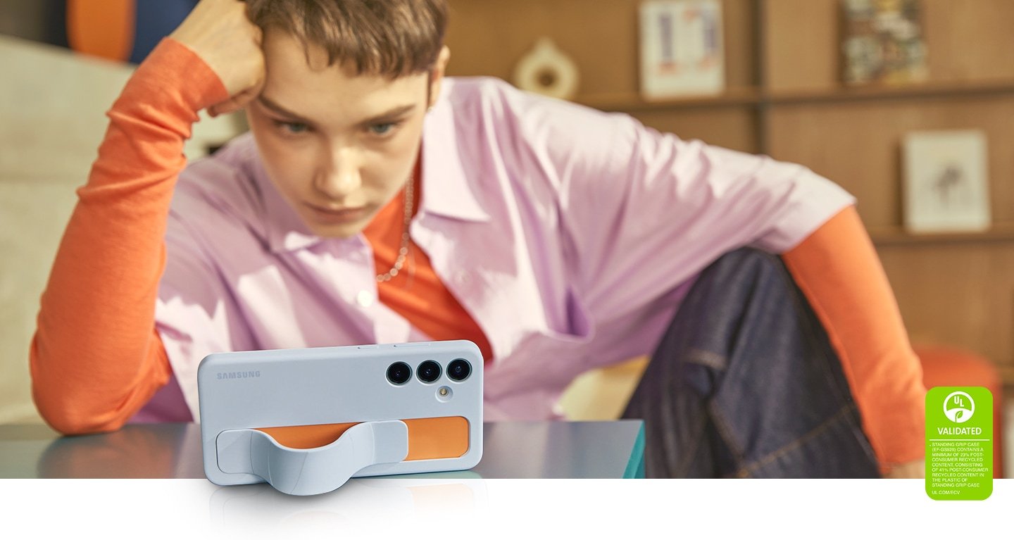 Une jeune étudiante soutient sa tête de la main tout en regardant l’écran de son Galaxy S24 Plus dans un étui ergonomique vertical Bleu Clair. La sangle forme un support qui aide le téléphone à se tenir debout, illustrant une situation pratique où la sangle est fixée pour visualiser confortablement l’écran. Le texte indique que L’ÉTUI ERGONOMIQUE VERTICAL CERTIFIÉ UL (EF-GS926) CONTIENT AU MOINS 23 % DE MATIÈRES RECYCLÉES POST-CONSOMMATION, AVEC 41 % DE MATIÈRES RECYCLÉES POST-CONSOMMATION DANS LE PLASTIQUE DE L’ÉTUI ERGONOMIQUE VERTICAL. UL.COM/ECV