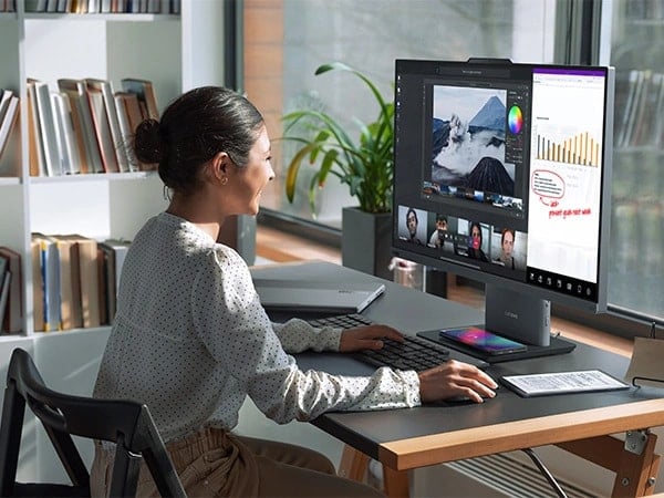 Lenovo ThinkCentre Neo 50a G5 27&Prime; all-in-one desktop PC &ndash; front right view, showing a woman working on a software using wireless keyboard and mouse, along with having a video conference and the base wirelessly charging a mobile phone in her home office setup.