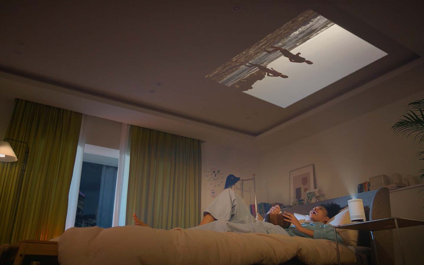 A woman is sitting on her bed eating popcorn, watching a video The Freestyle is projecting onto the wall in front of bed. Then she tilts The Freestyle so it points upwards and lays back with her head on the pillow, continuing to enjoying the video that&rsquo;s now projected onto the ceiling.