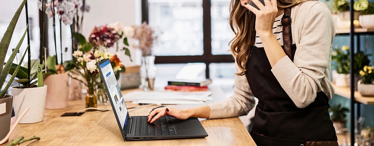 Une femme discute au téléphone avec son smartphone et tape avec sa main droite sur le PC portable Lenovo ThinkPad X1 Carbon Gen 12 qui se trouve sur une table entourée de plantes.