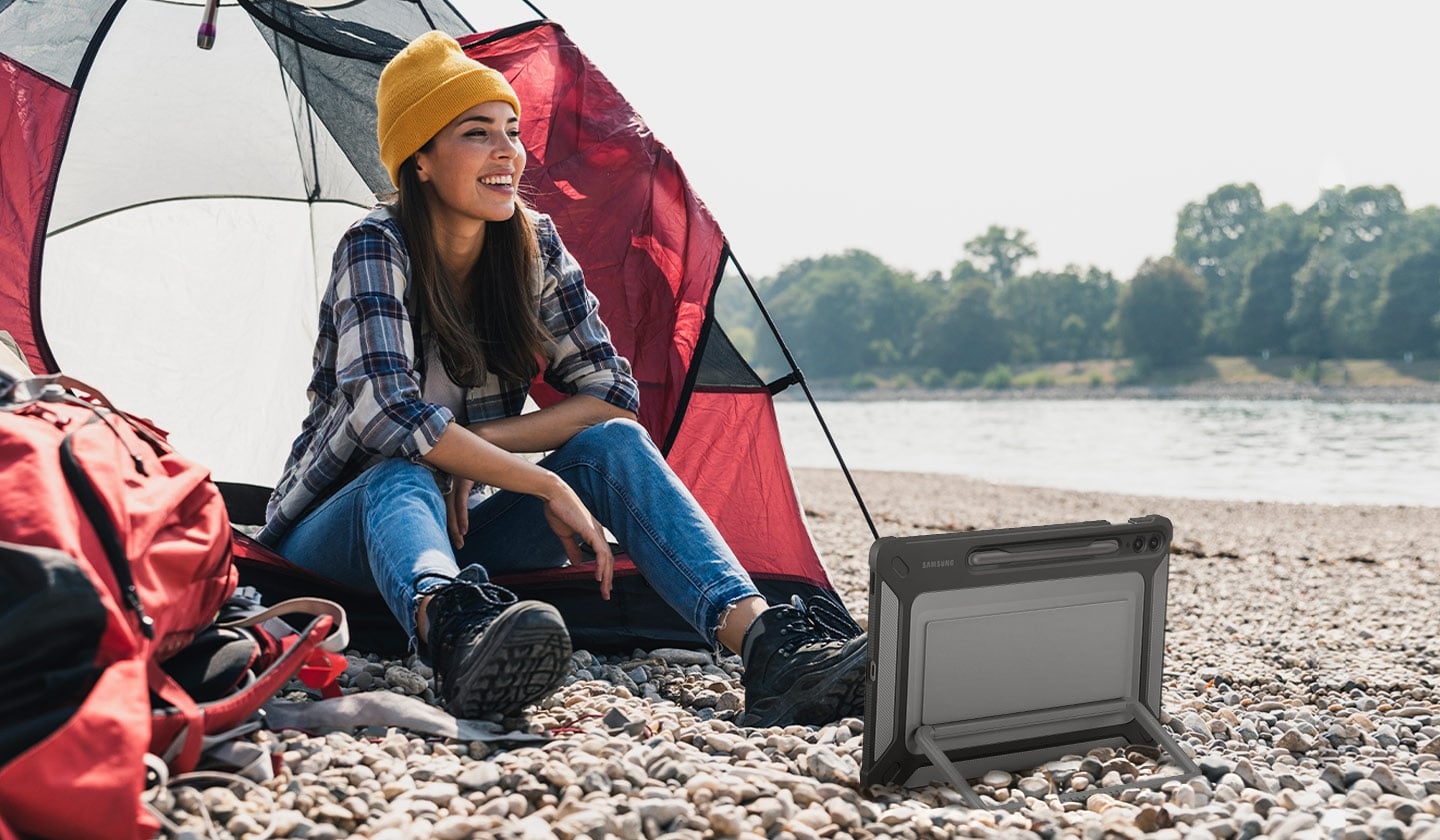 A woman sitting in front of a camping tent on a rocky surface is smiling. A Galaxy Tab S9 FE+ device with an Outdoor Cover on is placed horizontally in front of her propped up on a kickstand on the back of the cover.