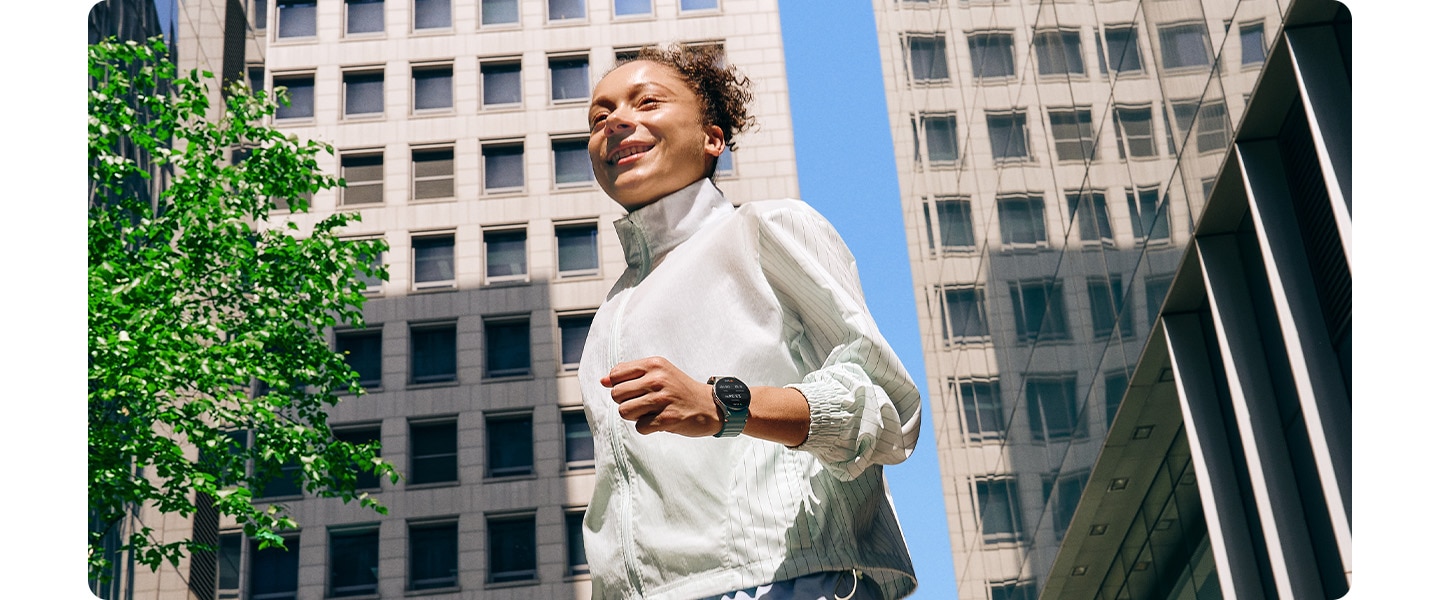 Une femme en tenue de sport portant une Galaxy Watch7 marche dans un quartier urbain entour&eacute; de b&acirc;timents.
