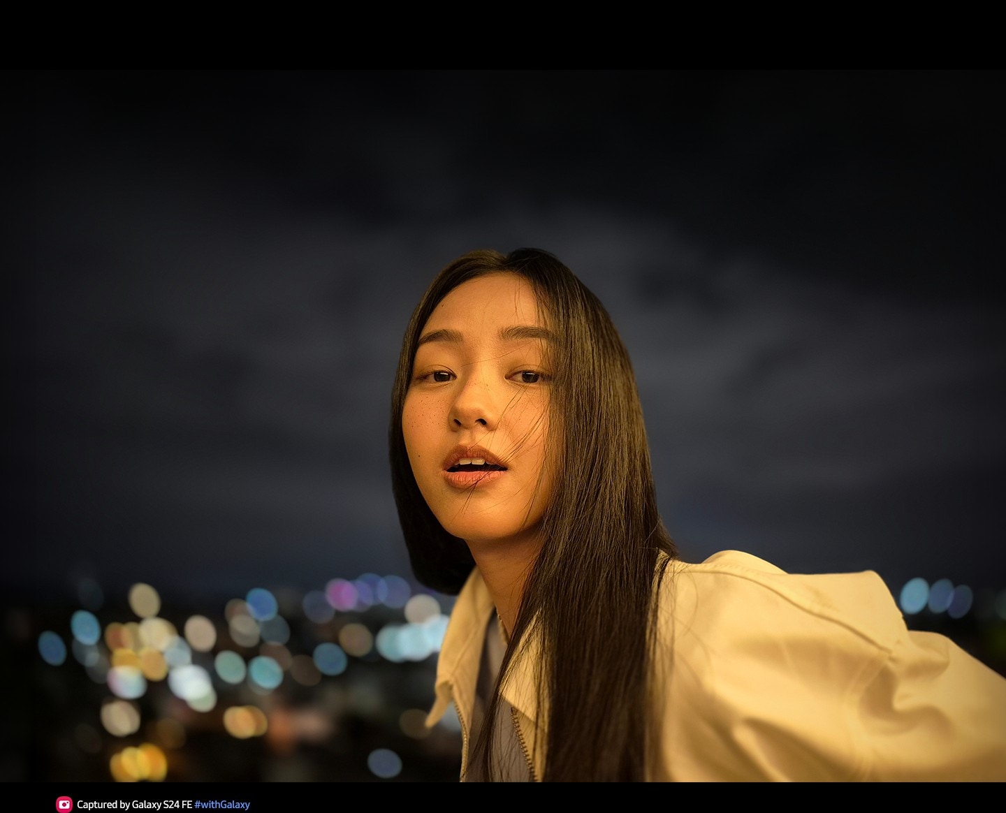 Photo d&rsquo;une femme en plein air la nuit avec un visage &eacute;clair&eacute; et des lumi&egrave;res de ville floues en arri&egrave;re-plan. Le texte indique Photo prise avec le Galaxy S24 FE #withGalaxy.