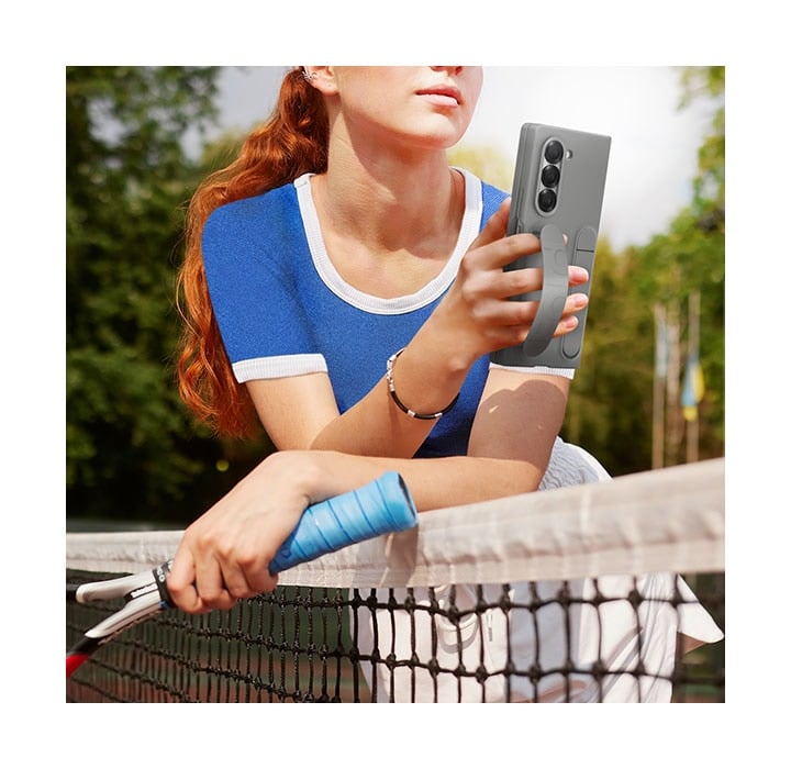 Une jeune femme sur un court de tennis s&rsquo;appuie sur le filet, tenant confortablement un Galaxy Z Fold6. Le t&eacute;l&eacute;phone est dans un &eacute;tui en silicone Gris et la femme tient le t&eacute;l&eacute;phone avec la poign&eacute;e de l&rsquo;&eacute;tui entre ses doigts.