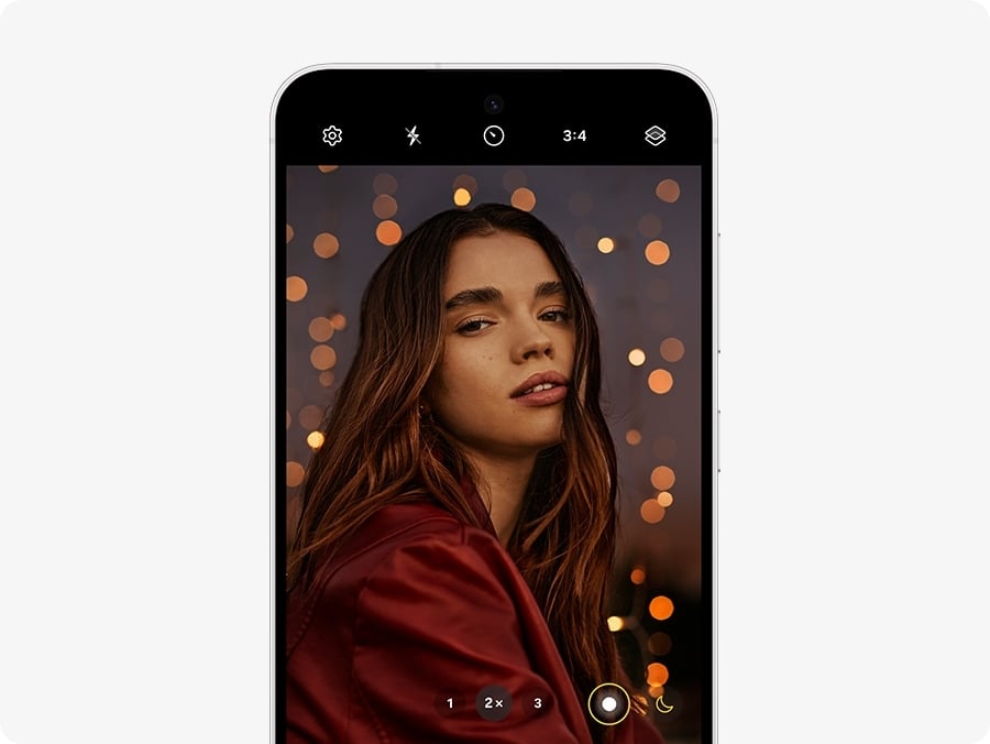 Un Galaxy S24 FE vu de face, affichant la photo d&rsquo;une femme souriante dans une pi&egrave;ce faiblement &eacute;clair&eacute;e. L&rsquo;&eacute;cran affiche l&rsquo;appareil photo en fonctionnement, r&eacute;gl&eacute; sur un zoom 2x avec un format d&rsquo;image 3:4.
