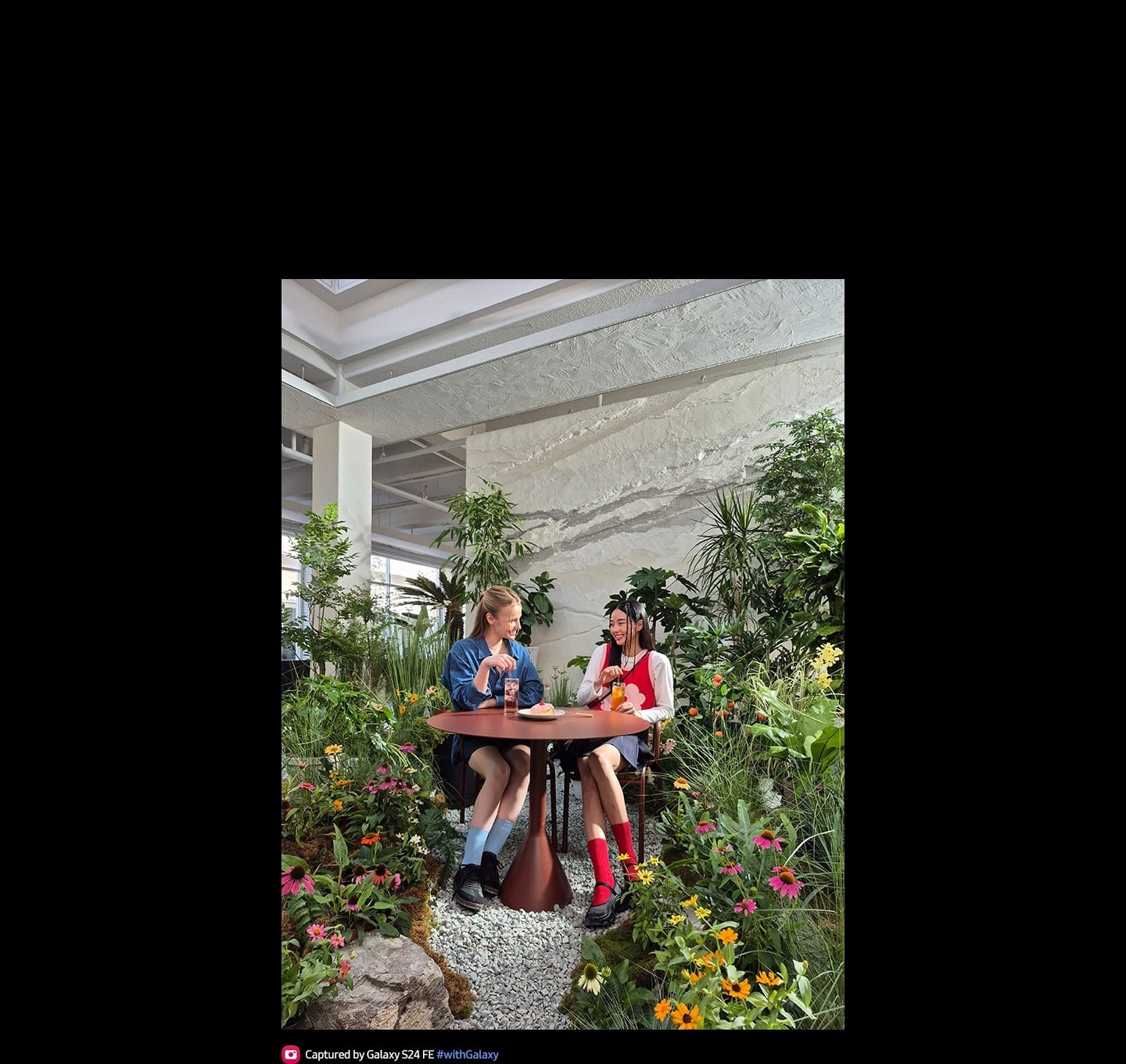 Photo de deux personnes assises ensemble &agrave; une table entour&eacute;e de plantes et de fleurs d&rsquo;int&eacute;rieur. Le texte indique Photo prise avec le Galaxy S24 FE #withGalaxy.