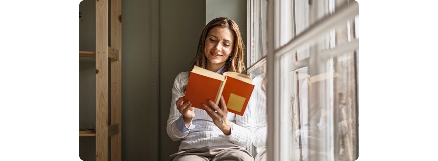 A woman sitting indoors, reading by a window. A line down the screen splits it into two sections, comparing a low-light shot on the left with a bright-light shot on the right.
