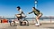 A split-screen with a line in the middle shows a man and a woman near the beach. On the left, which appears slightly blurry, the man is riding a bike. On the right side, which is sharp and clear, the woman is holding onto the man's bike wearing roller skates, highlighting the OIS feature of Galaxy A26 5G.