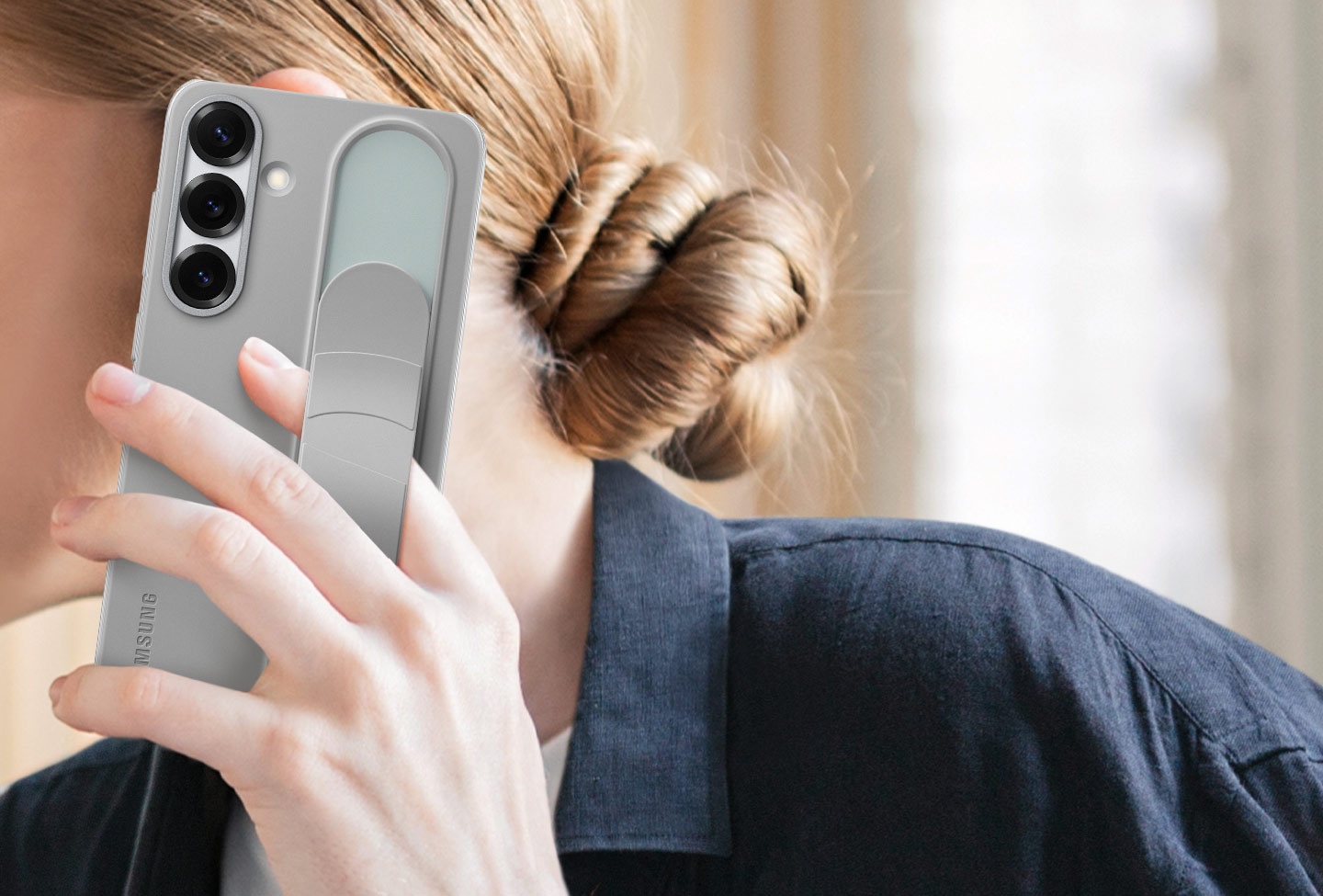 Une femme tient un Galaxy S25+ &eacute;quip&eacute; de la Standing Grip Case en Gris. Un doigt tenant le t&eacute;l&eacute;phone est plac&eacute; sous la poign&eacute;e r&eacute;glable.