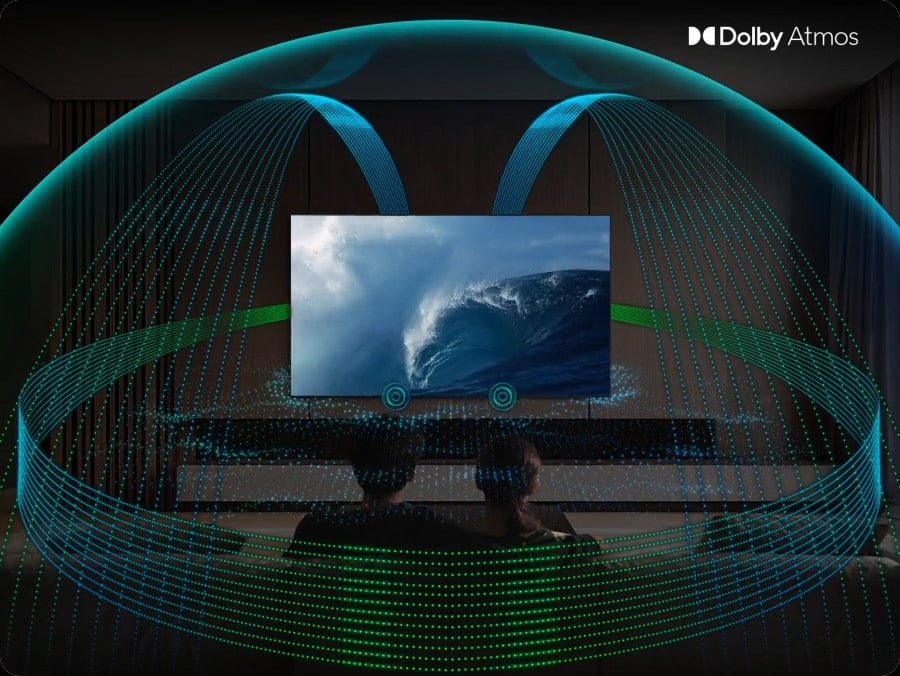 In a living room, a couple sits on a sofa and watches TV. The TV&rsquo;s front speakers are lit up and soundwaves emanate from a TV&rsquo;s side and up-firing speakers, travelling in multiple directions to surround the couple in a dome of sound and fill the room. Dolby Atmos logo.