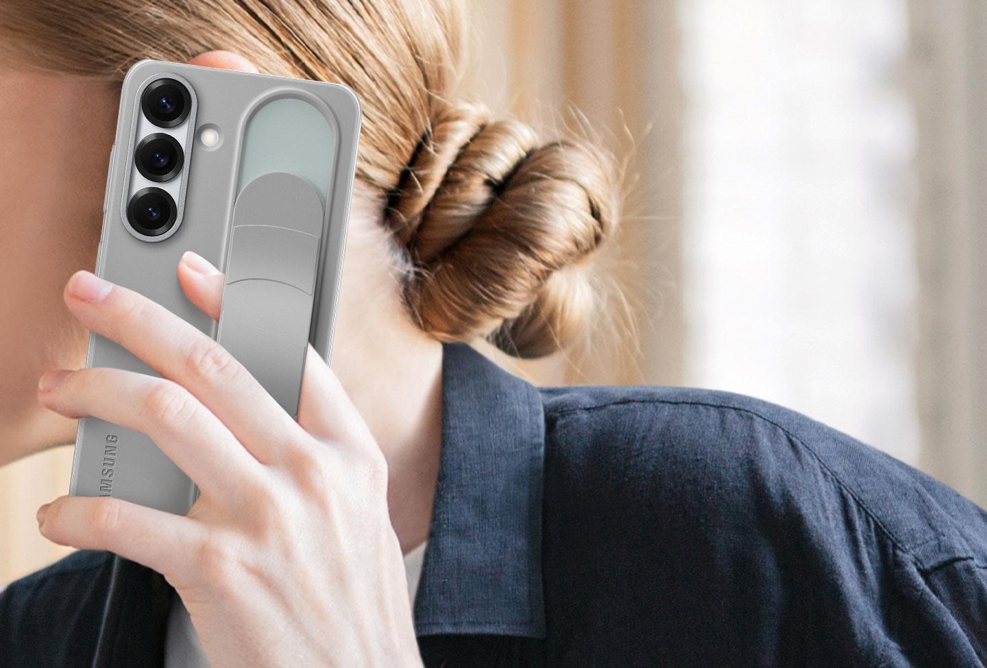 Une femme tient un Galaxy S25 &eacute;quip&eacute; de la Standing Grip Case en Gris. Un doigt tenant le t&eacute;l&eacute;phone est rang&eacute; sous la poign&eacute;e r&eacute;glable.