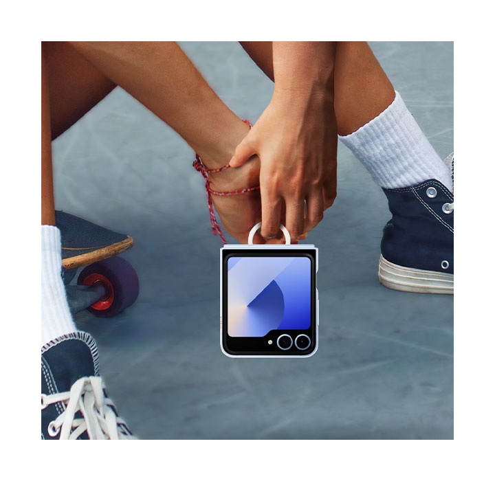 Un gros plan extrême d’une personne assise sur son skateboard tout en tenant confortablement son Galaxy Z Flip6 dans un étui transparent. La personne tient le téléphone uniquement avec son index à l’aide de l’anneau pour transport facile.