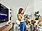 A woman sings into her phone, while the TV behind her displays karaoke lyrics. Her friends smile in the background.