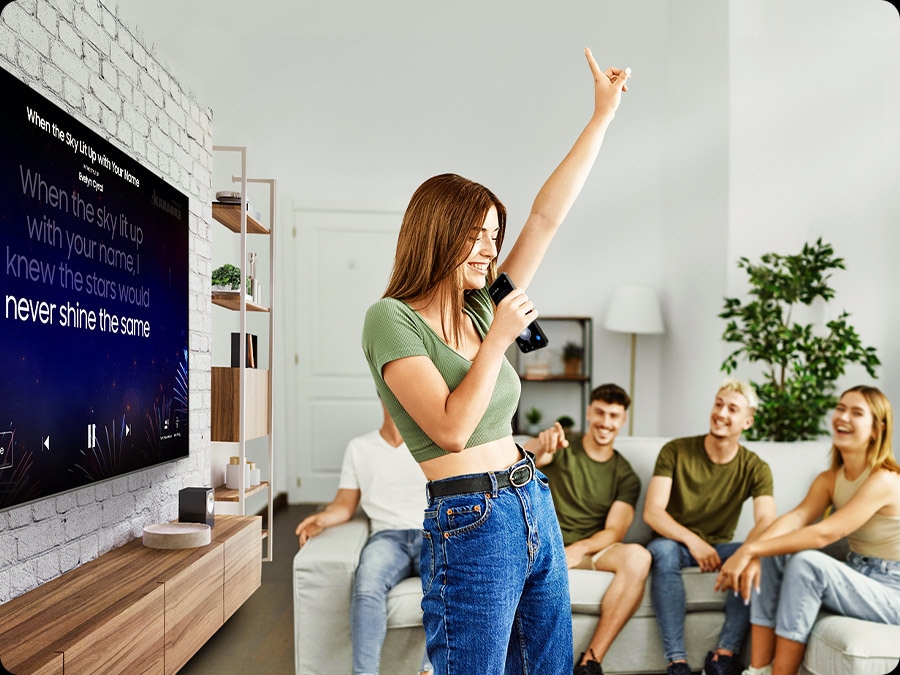 A woman sings into her phone, while the TV behind her displays karaoke lyrics. Her friends smile in the background.