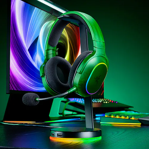 Vert gaming headset with a microphone, on a stand in front of a colorful monitor, with a keyboard behind, lit by RGB lights.