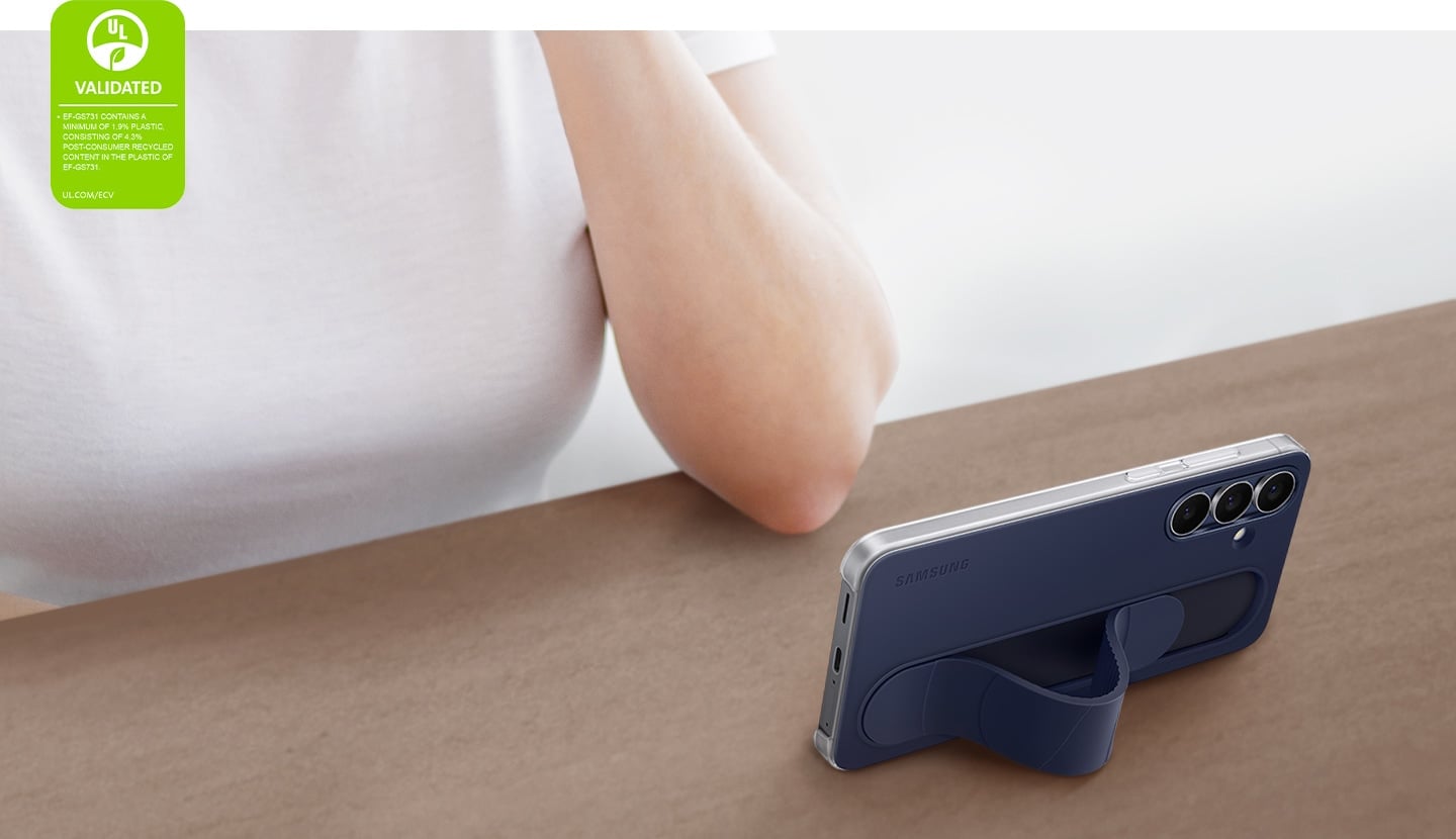 A Galaxy S25 FE encased in the Standing Grip Case in Dark Blue stands horizontally in a slanted angle on a table. The phone is being used by a person for viewing purposes. On the corner of the photo is a UL Environmental Claim Validation Mark that reads 'EF-GS731 CONTAINS A MINIMUM OF 1.9% PLASTIC, CONSISTING OF 4.3%POST-CONSUMER RECYCLED CONTENT IN THE PLASTIC OF EF-GS731. UL.COM/ECV'.