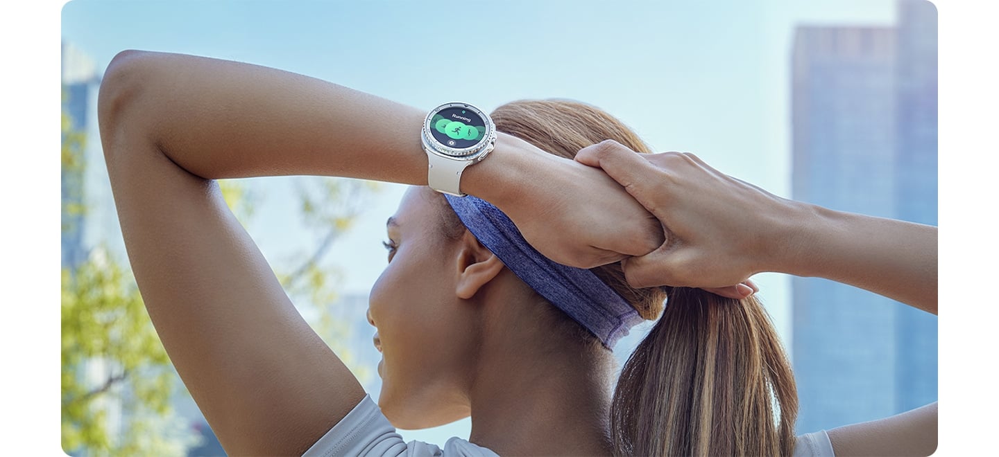 Une femme portant une Galaxy Watch8 Classic associ&eacute;e &agrave; un bracelet Sport Blanc pour Galaxy Watch8 &eacute;tire ses bras derri&egrave;re sa t&ecirc;te avant de commencer son entra&icirc;nement en ville.