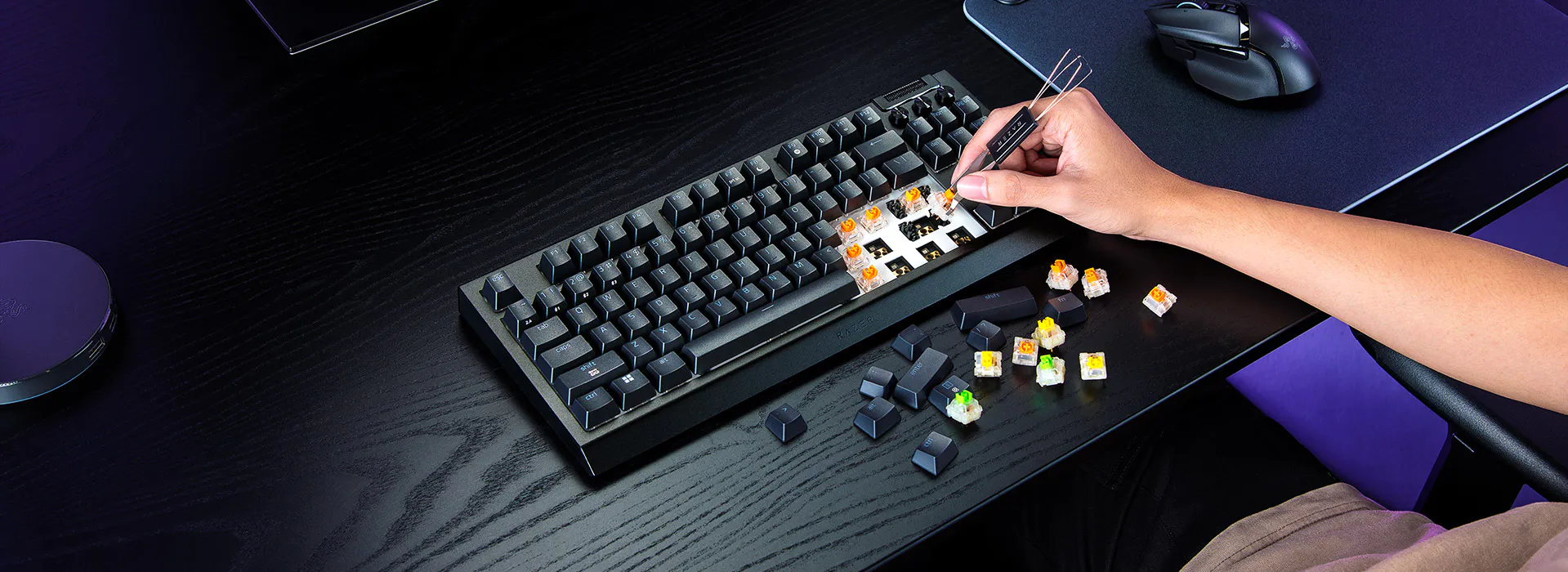 Une personne r&eacute;pare un clavier m&eacute;canique, &agrave; l&rsquo;aide d&rsquo;une pince &agrave; &eacute;piler, avec plusieurs interrupteurs et touches color&eacute;s &eacute;parpill&eacute;s sur un bureau noir.