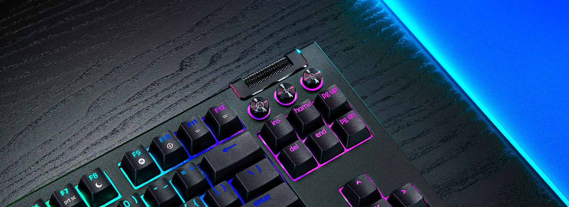 Clavier de jeu r&eacute;tro&eacute;clair&eacute; RVB avec molette de d&eacute;filement en m&eacute;tal et touches &eacute;tiquet&eacute;es F7 &agrave; F12, sur un bureau textur&eacute; sombre avec &eacute;clairage de bord LED bleu.