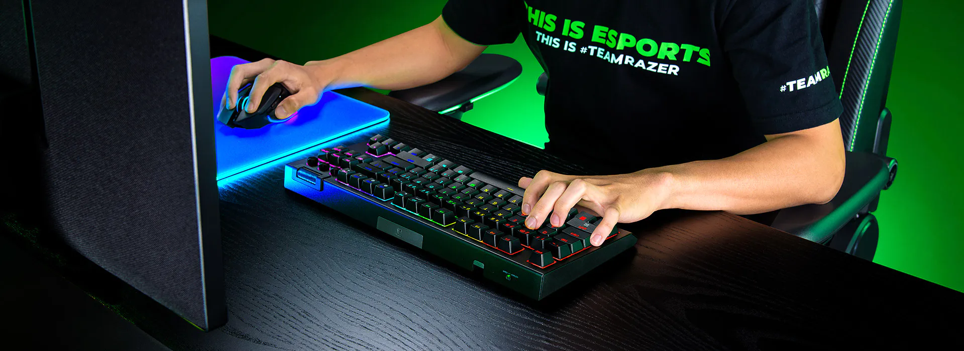 Personne utilisant un clavier, une souris et un moniteur de jeu dans un environnement sombre avec un &eacute;clairage vert et RVB ; portant un T-shirt noir avec du texte vert et blanc.