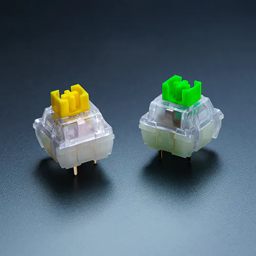 Deux commutateurs de clavier m&eacute;caniques transparents avec des tiges jaunes et vertes sur une surface sombre, montrant les m&eacute;canismes de contact internes.