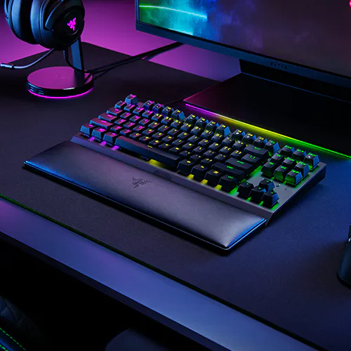 Clavier m&eacute;canique r&eacute;tro&eacute;clair&eacute; RVB avec repose-poignet sur un bureau de jeu sombre, un moniteur et des &eacute;couteurs avec &eacute;clairage violet.