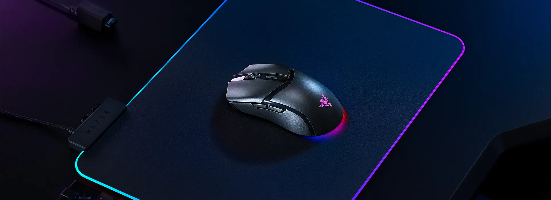 Une souris de jeu Razer sur un tapis de souris noir avec &eacute;clairage RVB, connect&eacute;e via un c&acirc;ble, avec un clavier filaire partiellement visible en dessous.