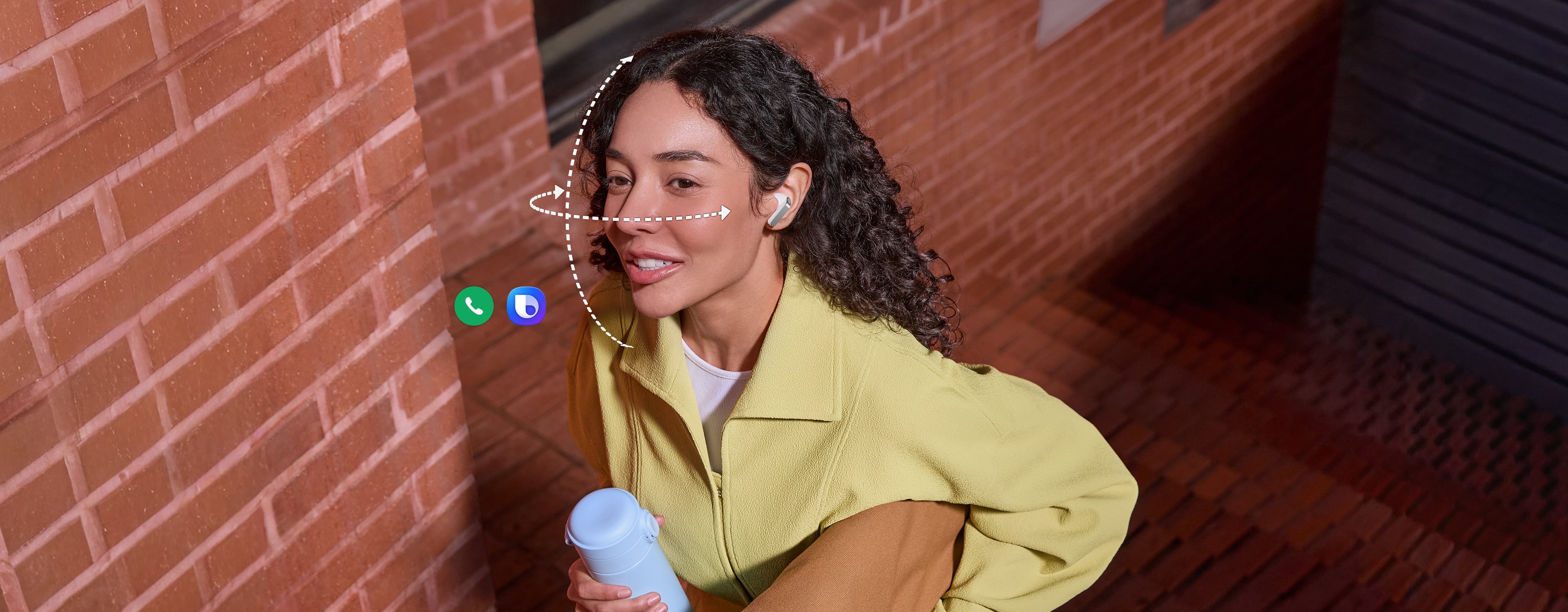 Une personne porte des Galaxy Buds4 Pro de couleur blanche, tout en tenant un gobelet dans une main et un sac dans l’autre. Des lignes verticales et horizontales en pointillés s’enroulent autour de son visage pour illustrer la fonction Geste de la tête disponible sur les Galaxy Buds4 Pro. L’icône de l’application d’appel et l’icône Bixby indiquent que les deux fonctionnalités peuvent être utilisées pour le geste de la tête.