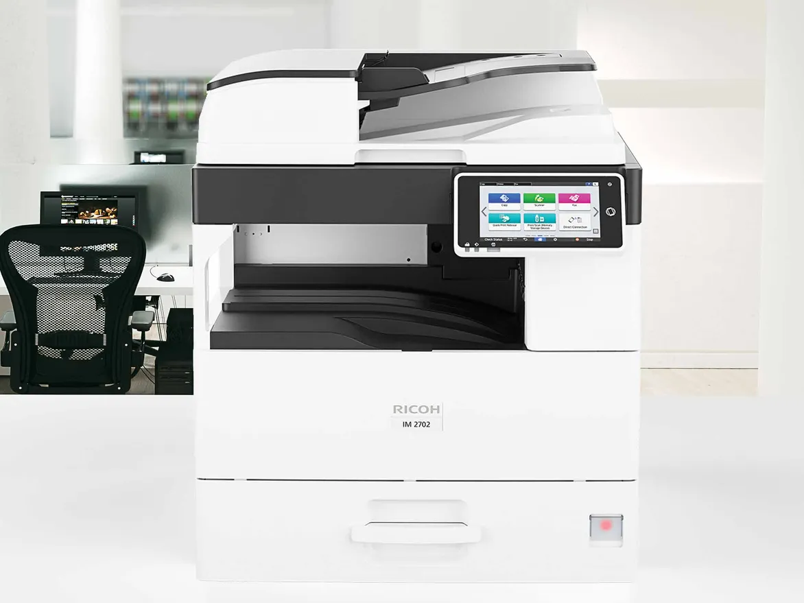 الطابعة متعددة الوظائف Ricoh IM 2702 من الأمام في مكتب مفتوح، وحدة تغذية المستندات مغلقة وشاشة اللمس تعرض وظائف النسخ والماسح الضوئي والفاكس
