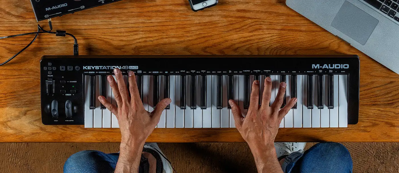 Deux mains sur le Keystation 49 MK3 posé sur un bureau en bois, à côté d'un ordinateur portable et d'une interface audio