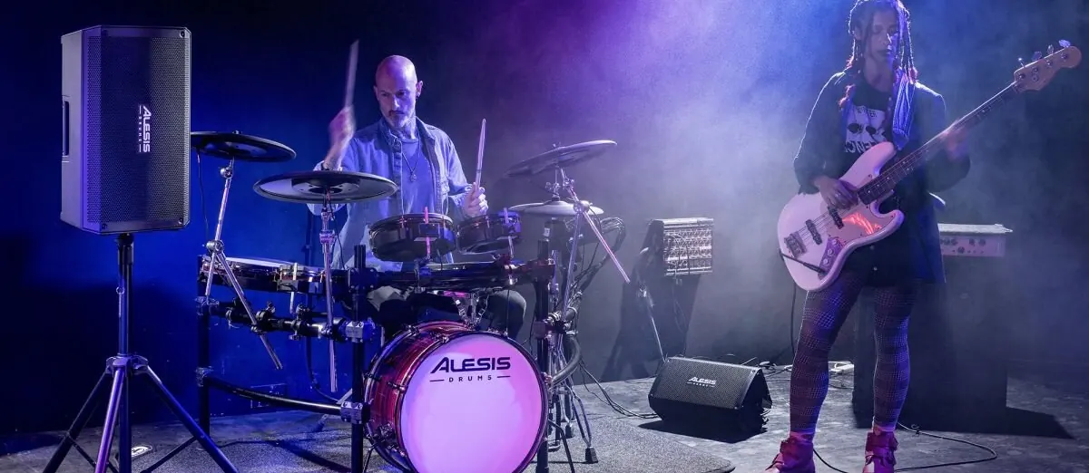 Batteur sur une batterie électronique Alesis en concert, une Strike Amp 12 MK2 sur pied derrière lui et un bassiste à côté